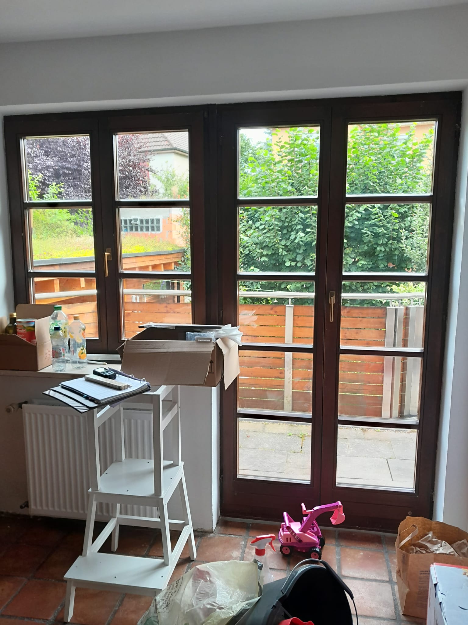 Helles Zimmer mit brauner Terrassentür, Umzugskartons, weißer Trittleiter und rosa Spielzeug